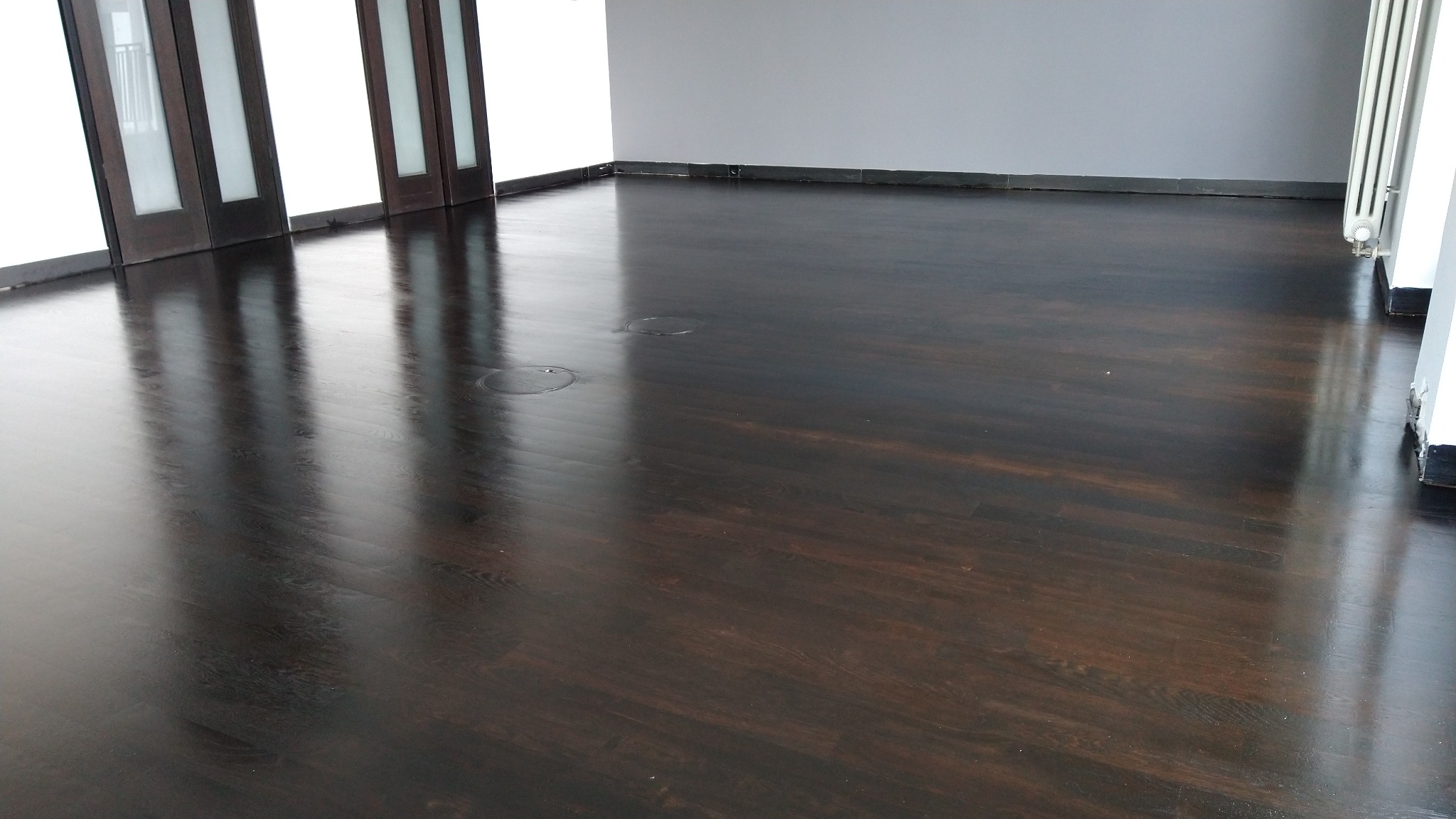 Błyszcząca, ciemna drewniana podłoga z widocznymi słojami, odnowiona w pustym pokoju z białymi ścianami i ciemnymi drzwiami balkonowymi.