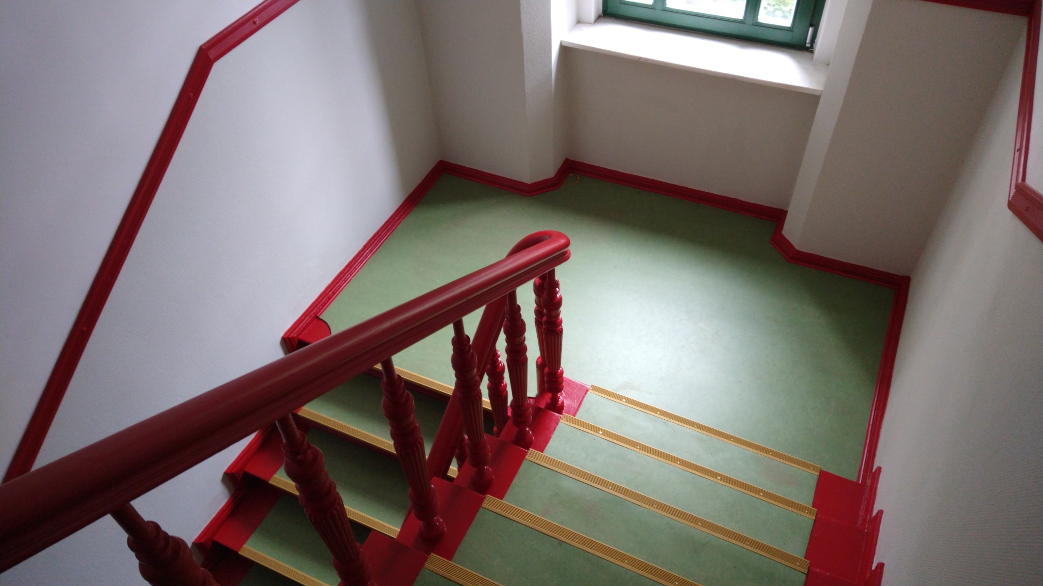 Widok z góry na odnowione schody z zieloną posadzką, czerwonymi balustradami i listwami, oraz złotymi paskami antypoślizgowymi na stopniach, w pobliżu okna.