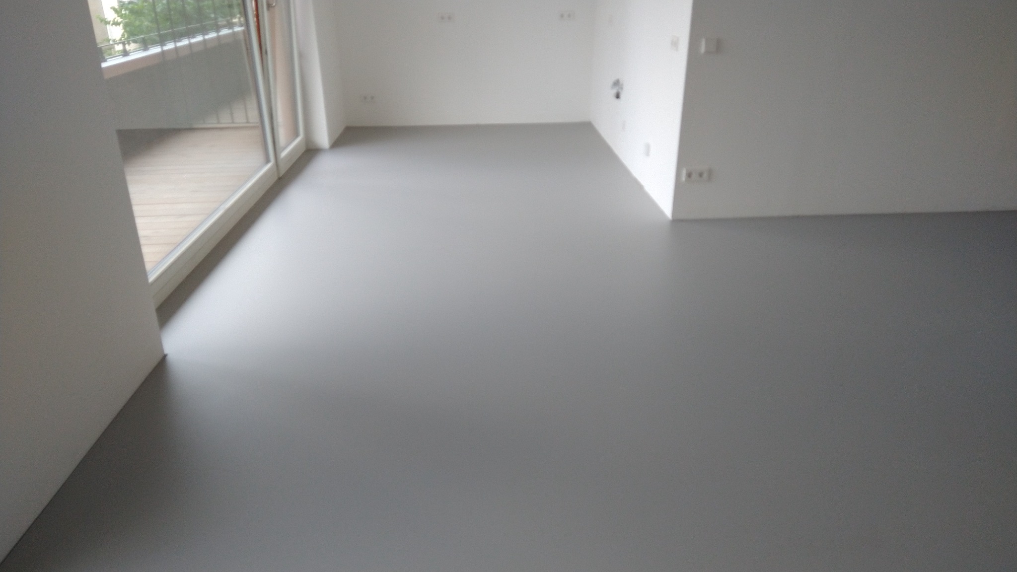 Szara, gładka podłoga żywiczna w jasnym, minimalistycznym wnętrzu z balkonem i białymi ścianami.