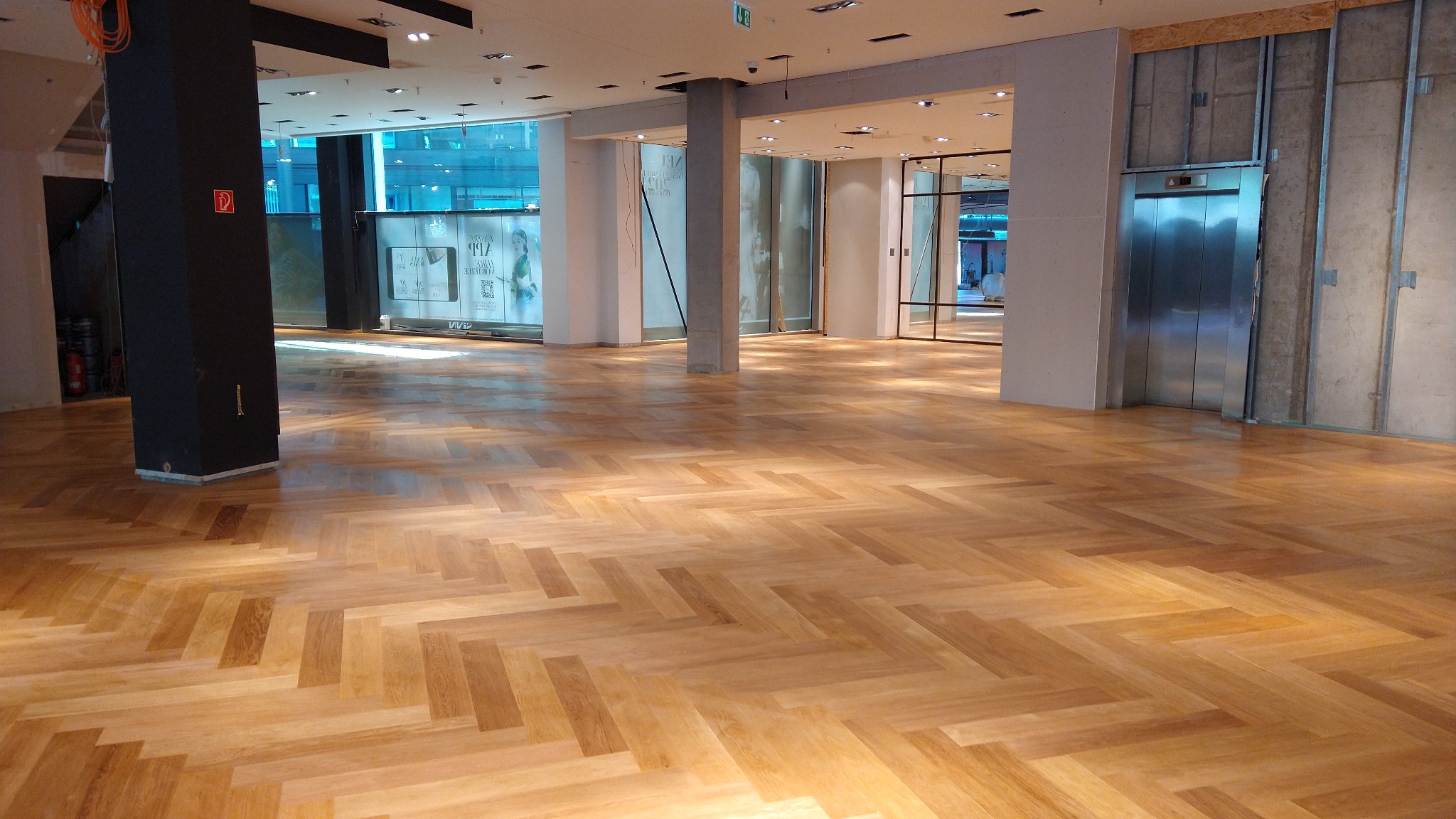 Jasna, lakierowana podłoga z parkietu ułożonego w jodełkę francuską w przestronnym, nowoczesnym wnętrzu biurowym z dużymi oknami i kolumnami.