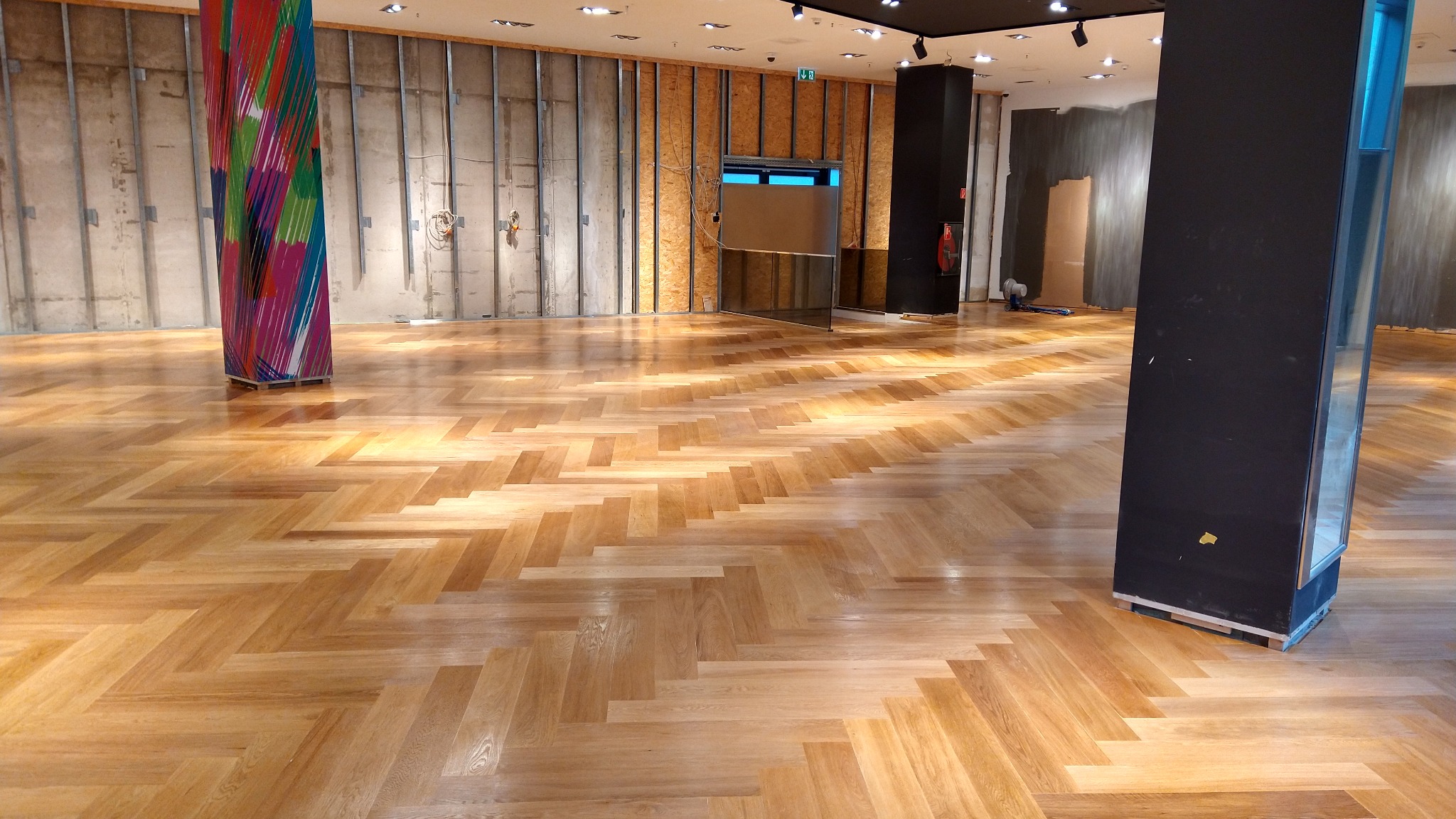 Jasny, dębowy parkiet ułożony we wzór jodełki francuskiej w dużym pomieszczeniu w trakcie remontu, widoczne surowe ściany i filary.