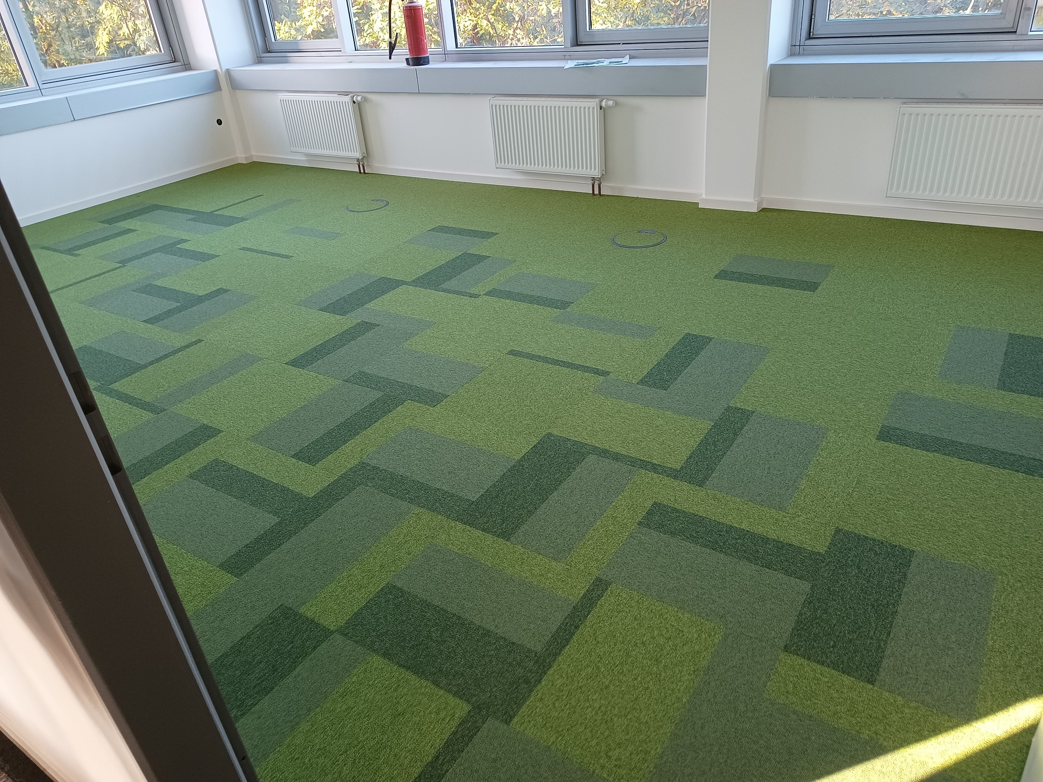 Wnętrze biurowe z nową wykładziną dywanową w odcieniach zieleni, ułożoną w geometryczny wzór. Widoczne okna z białymi ramami, grzejniki i gniazda podłogowe.