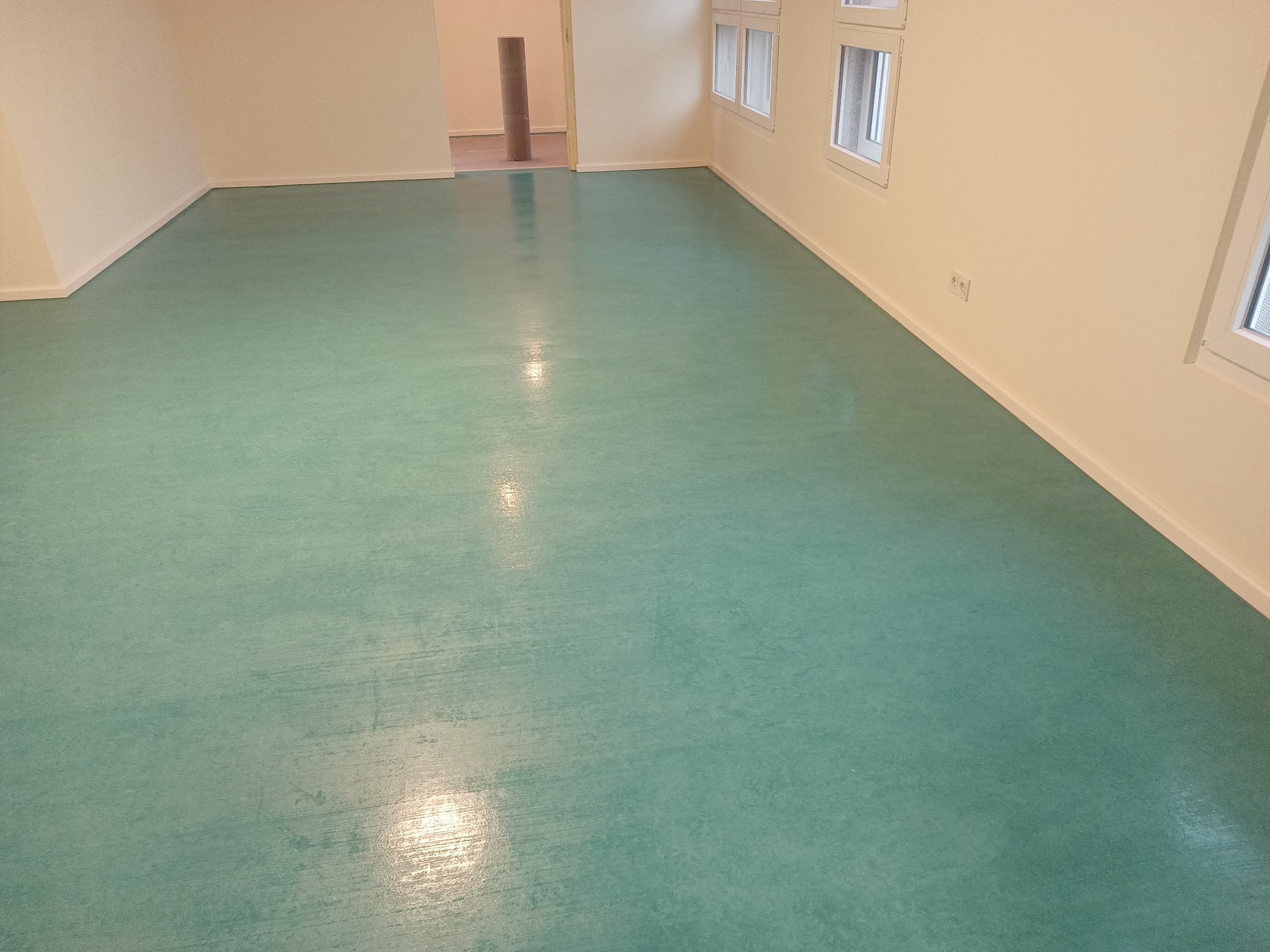 Jasnozielona, gładka podłoga w pustym pokoju z białymi ścianami i kilkoma oknami. W tle widoczny zwinięty materiał podłogowy.