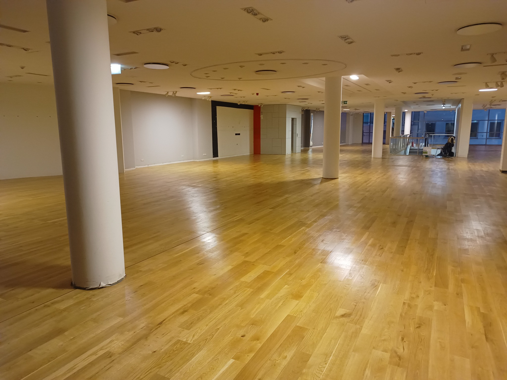 Duża, jasna sala z parkietem ułożonym we wzór jodełki, widoczne białe kolumny podtrzymujące sufit z okrągłymi oprawami oświetleniowymi. W tle osoba pracująca przy oknie.