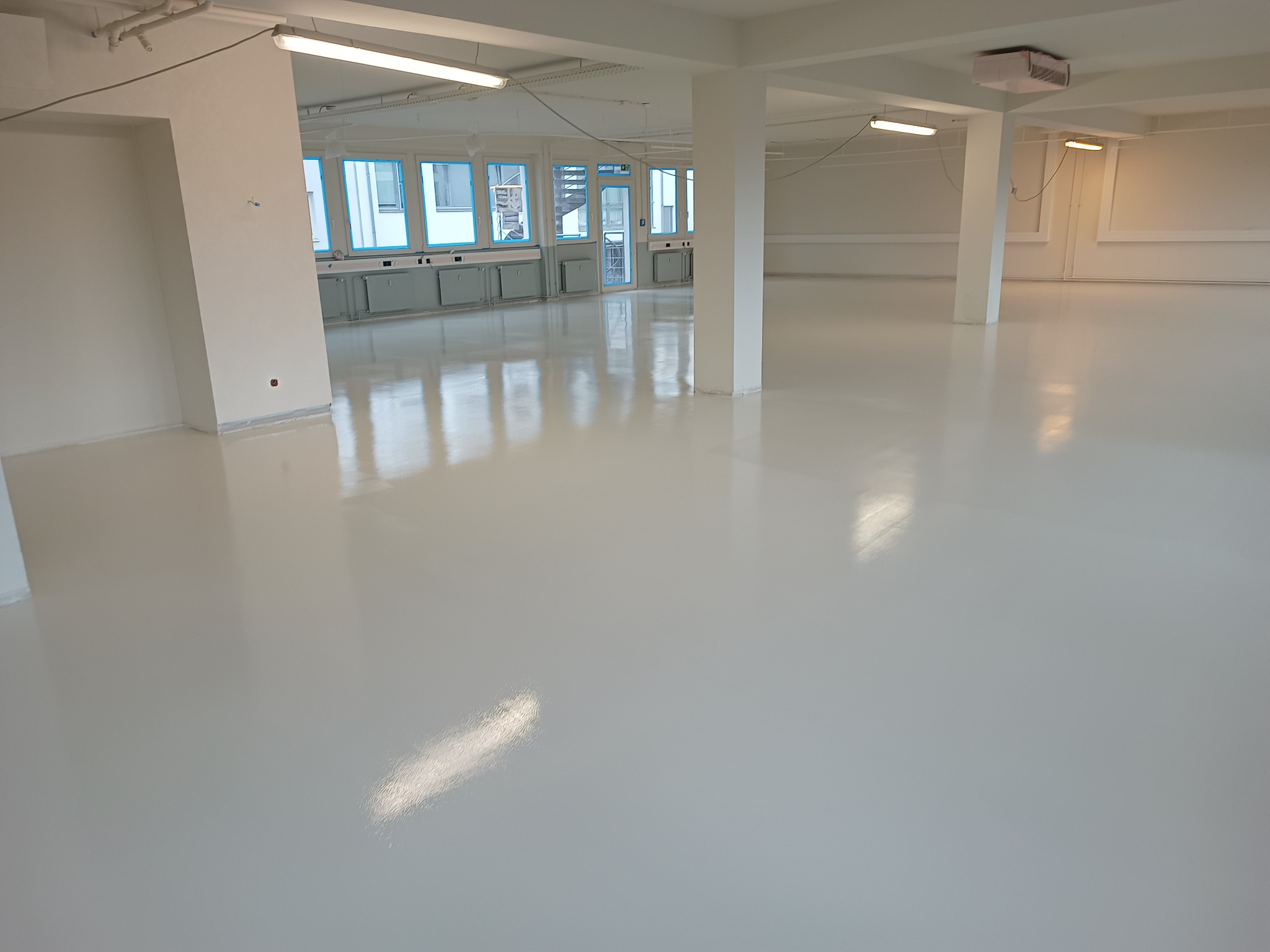 Jasna, błyszcząca posadzka żywiczna w przestronnym, industrialnym wnętrzu z kolumnami i dużymi oknami.