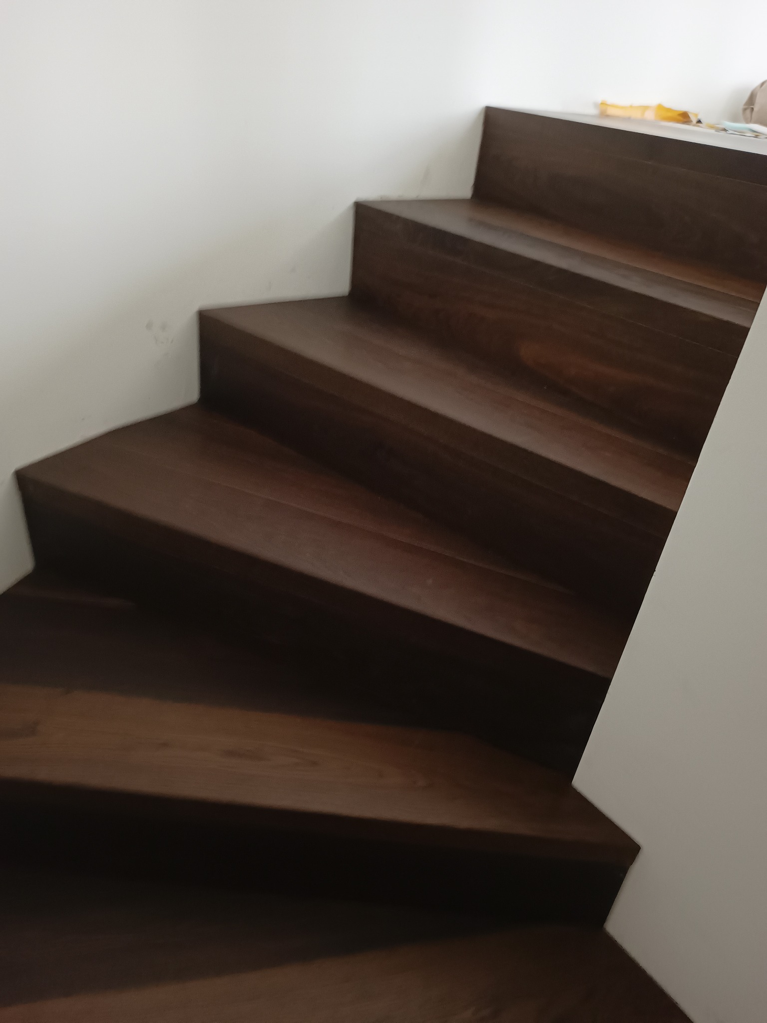 Ciemne, drewniane schody z widocznym usłojeniem, przylegające do białej ściany, sfotografowane z dołu.