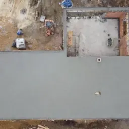 Widok z góry na wylewkę betonową fundamentu domu, z elementami szalunku, częściowo wymurowanymi ścianami z cegły, schodami wejściowymi i przygotowanym terenem pod budowę.