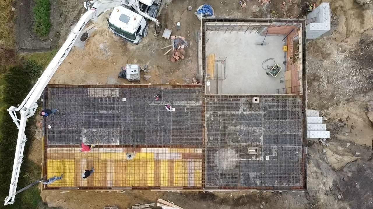Widok z góry na plac budowy z betonowym fundamentem domu, zbrojenie ułożone, pracownicy przy pompie do betonu, obok widoczny wykop pod schody.
