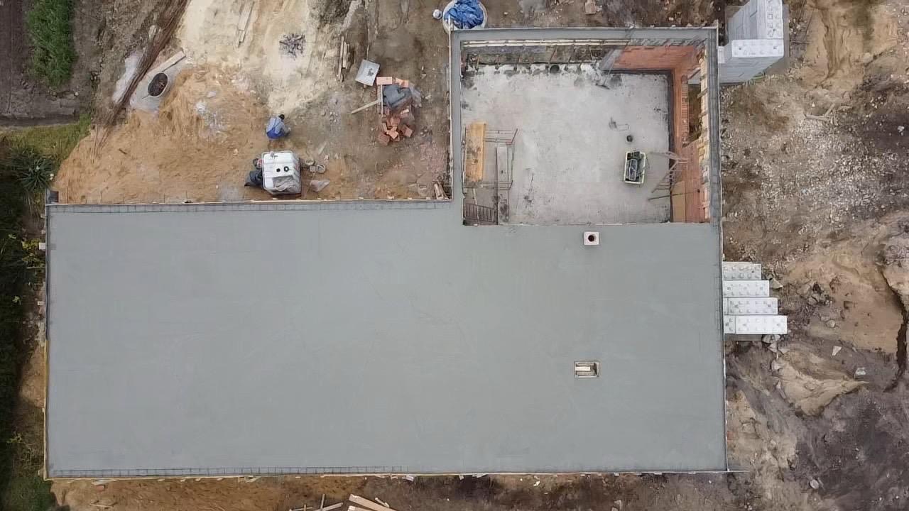 Widok z góry na wylewkę betonową fundamentu domu, z elementami szalunku, częściowo wymurowanymi ścianami z cegły, schodami wejściowymi i przygotowanym terenem pod budowę.