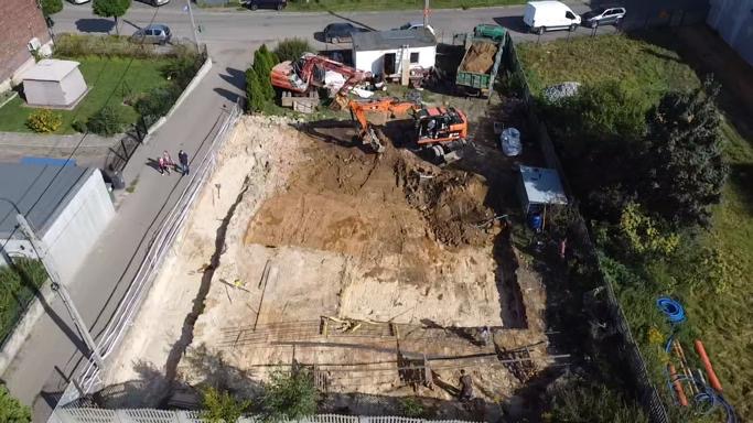 Widok z góry na plac budowy z wykopem pod fundamenty, dwie koparki, wywrotka z ziemią, widoczne zbrojenia, obok chodnik z przechodniami.
