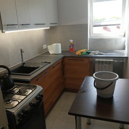 Czysto W Raz Dwa Trzy Daniel Szeksztełło - Umyta kuchnia z białymi szafkami, brązowymi frontami dolnych szafek, czarnym zlewem i kuchenką gazową; na blacie płyn do czyszczenia, ręcznik papierowy i ściereczki; na stole stoi wiadro z wodą.