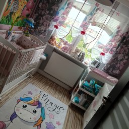 Czysto W Raz Dwa Trzy Daniel Szeksztełło - Umeblowany pokój dziecięcy z łóżeczkiem, regałem na pieluszki i dywanem z jednorożcem na podłodze z paneli, dekoracje okienne z motywem Myszki Miki i Wieży Eiffla.