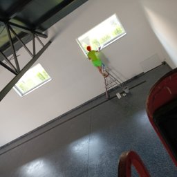 Czysto W Raz Dwa Trzy Daniel Szeksztełło - Osoba w jaskrawym stroju na drabinie czyści okno dachowe w przestronnym, nowoczesnym wnętrzu z widoczną konstrukcją dachu i elementami wyposażenia.