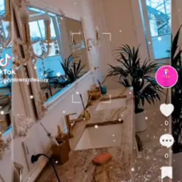 Luksusowa łazienka z podw&oacute;jną umywalką z granitu, złotymi bateriami i akcesoriami, ozdobiona roślinami doniczkowymi i lustrem w bogato zdobionej ramie; na blacie leży pomarańczowa gąbka.