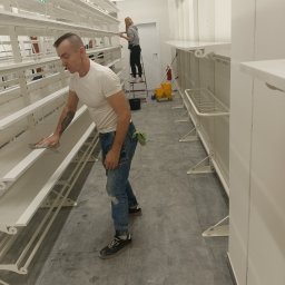 Czysto W Raz Dwa Trzy Daniel Szeksztełło - Pracownik w białej koszulce i dżinsach czyści półki w sklepie przy użyciu szmatki; w tle druga osoba na drabinie i sprzęt do sprzątania.