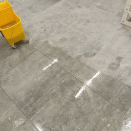 Czysto W Raz Dwa Trzy Daniel Szeksztełło - Szare płytki podłogowe częściowo umyte, z widocznymi smugami wody i żółtym wiadrem na kółkach w lewym górnym rogu, obok rozłożonej suszarki do ubrań.