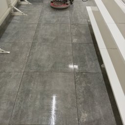 Czysto W Raz Dwa Trzy Daniel Szeksztełło - Automat do czyszczenia podłóg na szarych, kwadratowych płytkach, widoczny błyszczący ślad po czyszczeniu.