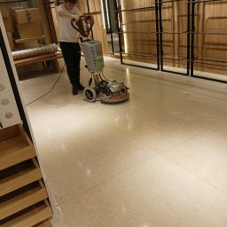 Czysto W Raz Dwa Trzy Daniel Szeksztełło - Pracownik w białej koszulce czyści jasną, kamienną podłogę w sklepie odzieżowym za pomocą profesjonalnej maszyny czyszczącej, widoczne mokre ślady po czyszczeniu.