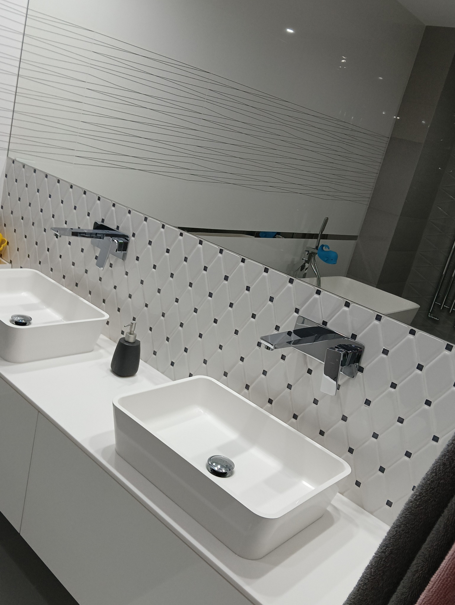 Nowoczesna łazienka z dwoma prostokątnymi umywalkami nablatowymi, chromowanymi bateriami i dozownikiem mydła na białym blacie, ozdobiona biało-czarną mozaiką na ścianie i dużym lustrem.