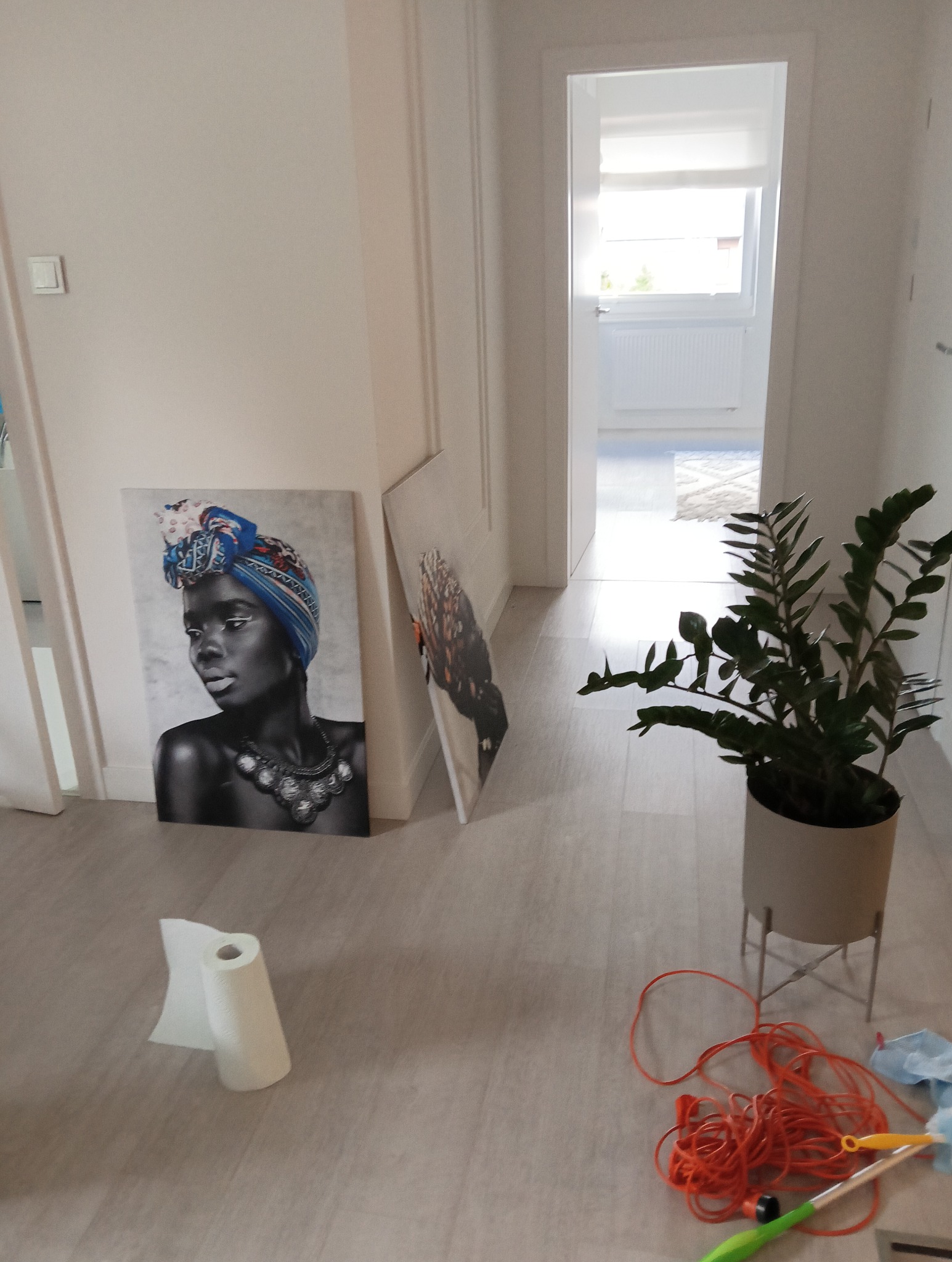 Korytarz w trakcie porządkowania: dwa obrazy oparte o ścianę, zwinięty ręcznik papierowy, doniczka z zieloną rośliną, pomarańczowy sznur i ściereczka do kurzu.