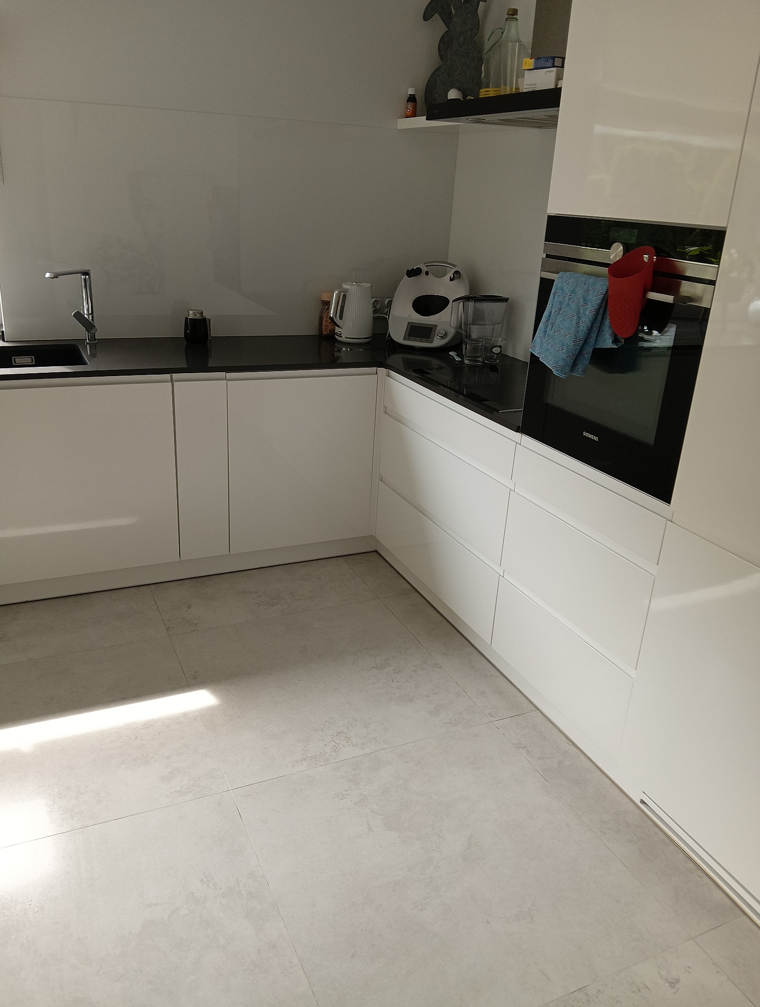 Nowoczesna, jasna kuchnia z białymi szafkami, czarnym blatem i sprzętami AGD, widoczne akcesoria kuchenne i tekstylia.