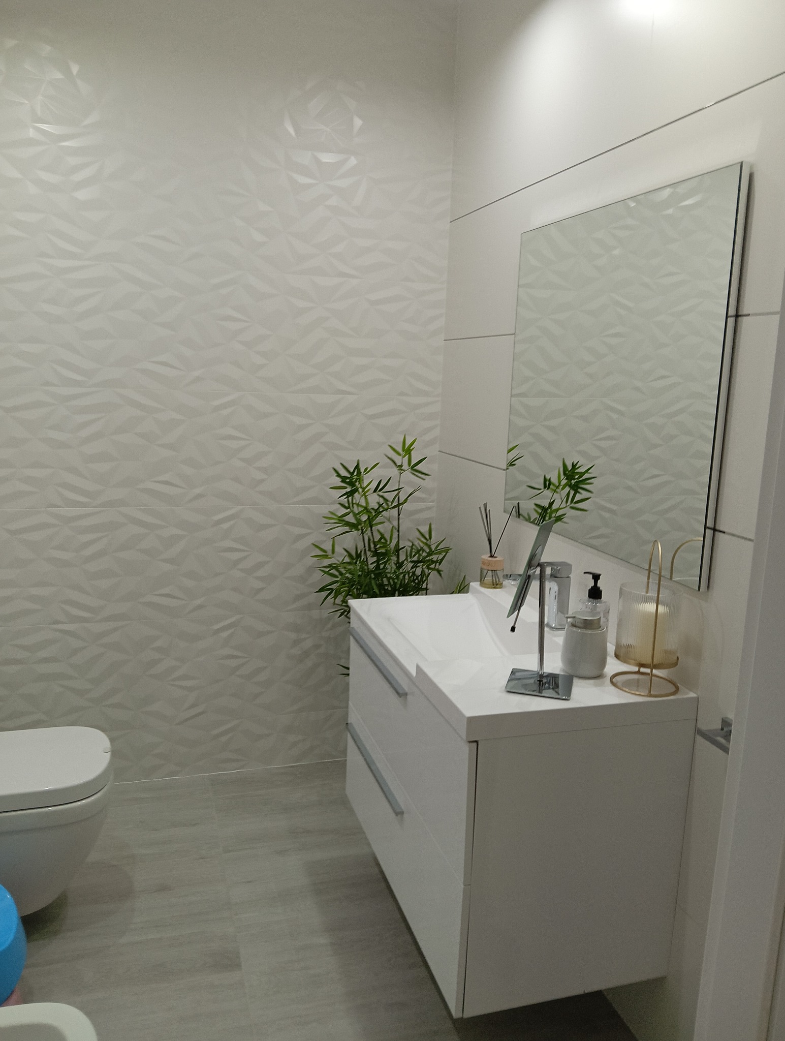 Nowoczesna łazienka w odcieniach bieli z umywalką, lustrem, dekoracyjną ścianą 3D, bambusem w doniczce i akcesoriami łazienkowymi.