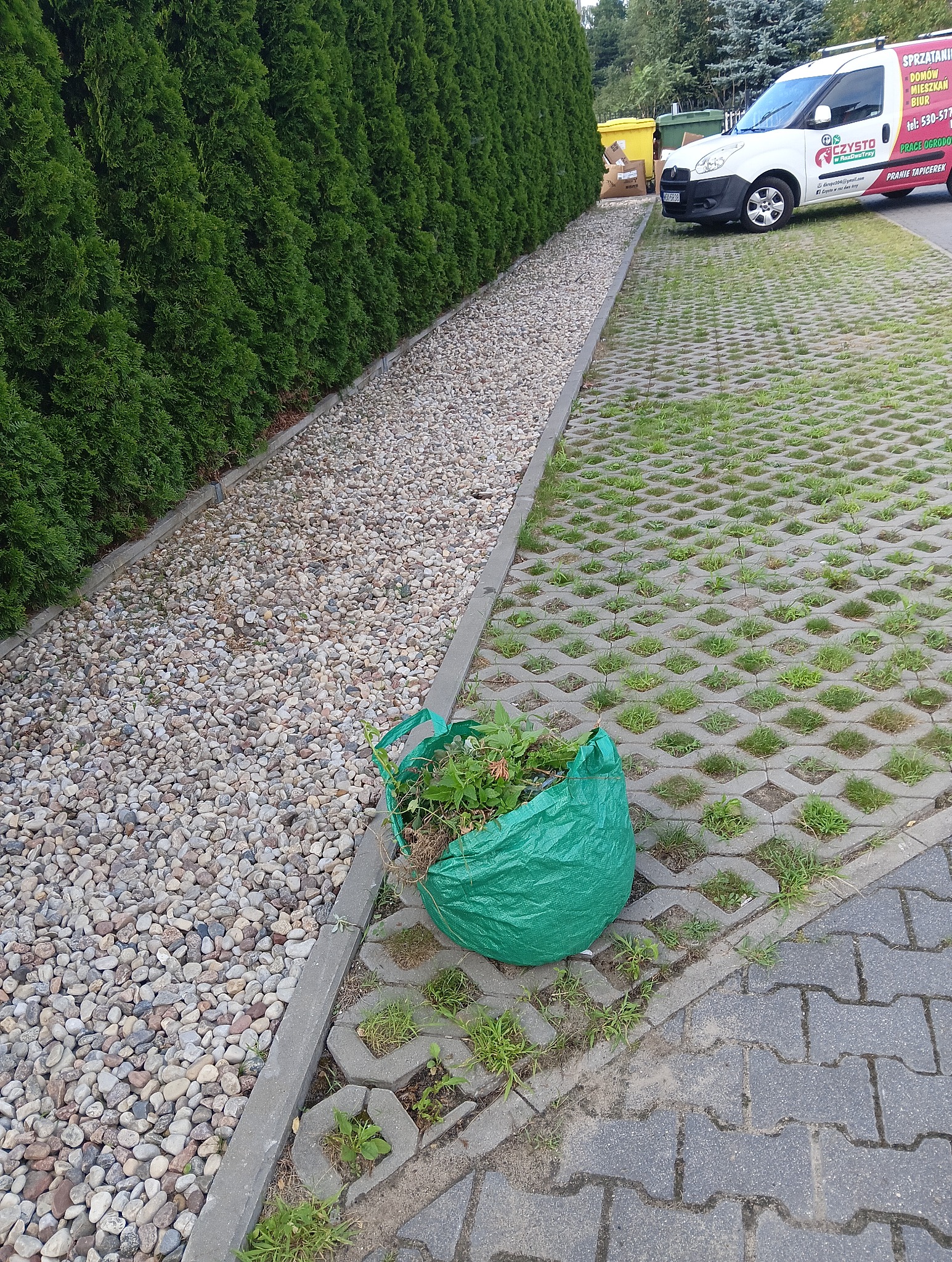 Zielony worek z resztkami roślin leży na betonowej ażurowej nawierzchni, obok rzędu tuj i wysypanych kamyków, w tle biały samochód dostawczy z reklamą usług porządkowych.