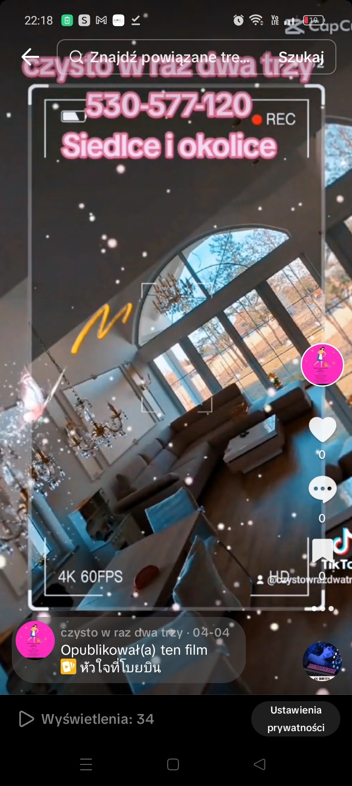 Wnętrze salonu z narożnikiem, widok z góry, duże okna z widokiem na drzewa, ozdobny żyrandol, nakładka wideo z numerem telefonu i nazwą miejscowości.