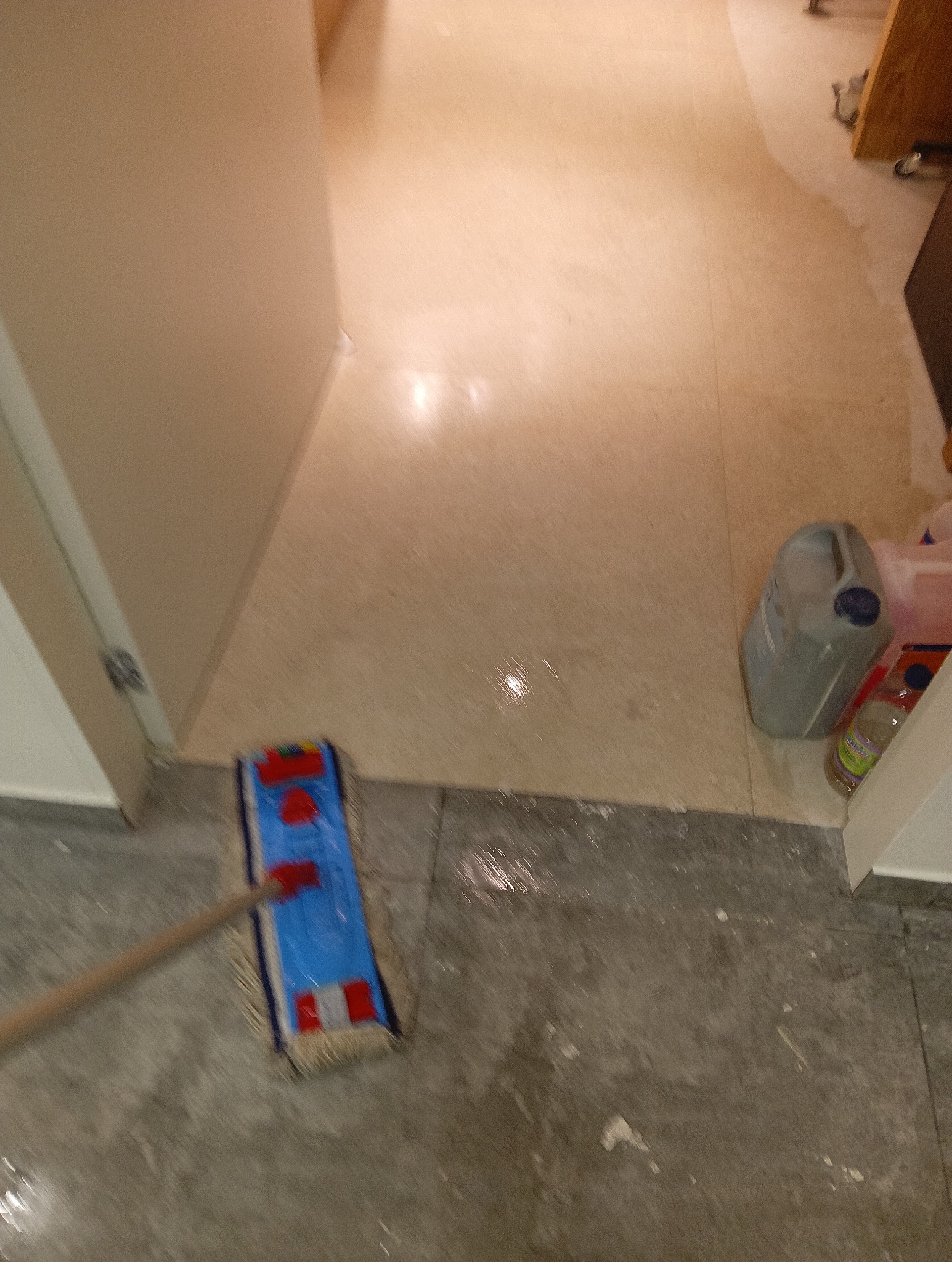 Mop z niebieską nakładką czyszczącą, przesuwany po mokrej, szarej podłodze z widocznymi smugami wody, obok ściany i pojemnika z płynem do mycia podłóg.