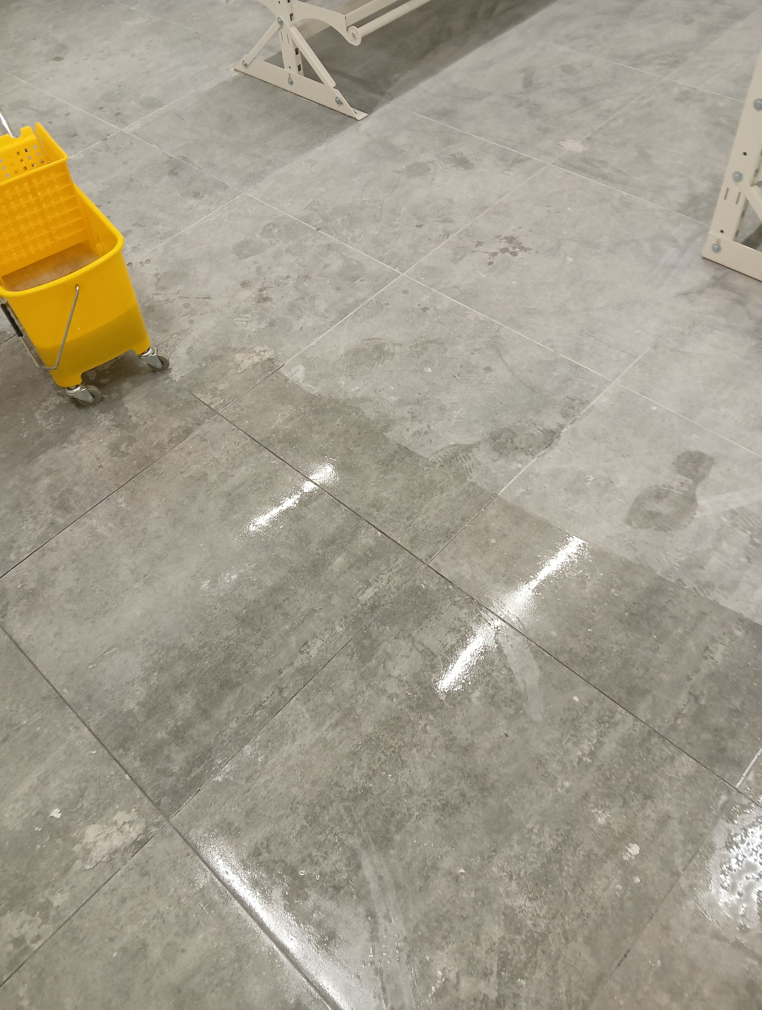 Szare płytki podłogowe częściowo umyte, z widocznymi smugami wody i żółtym wiadrem na kółkach w lewym górnym rogu, obok rozłożonej suszarki do ubrań.
