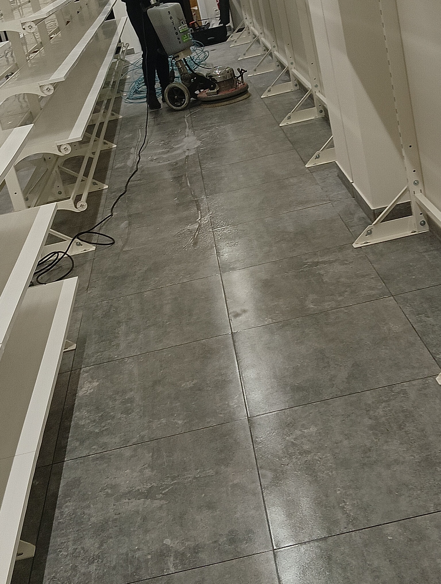 Czyszczenie podłogi z płytek w pomieszczeniu komercyjnym za pomocą szorowarki, widoczne białe regały w tle.