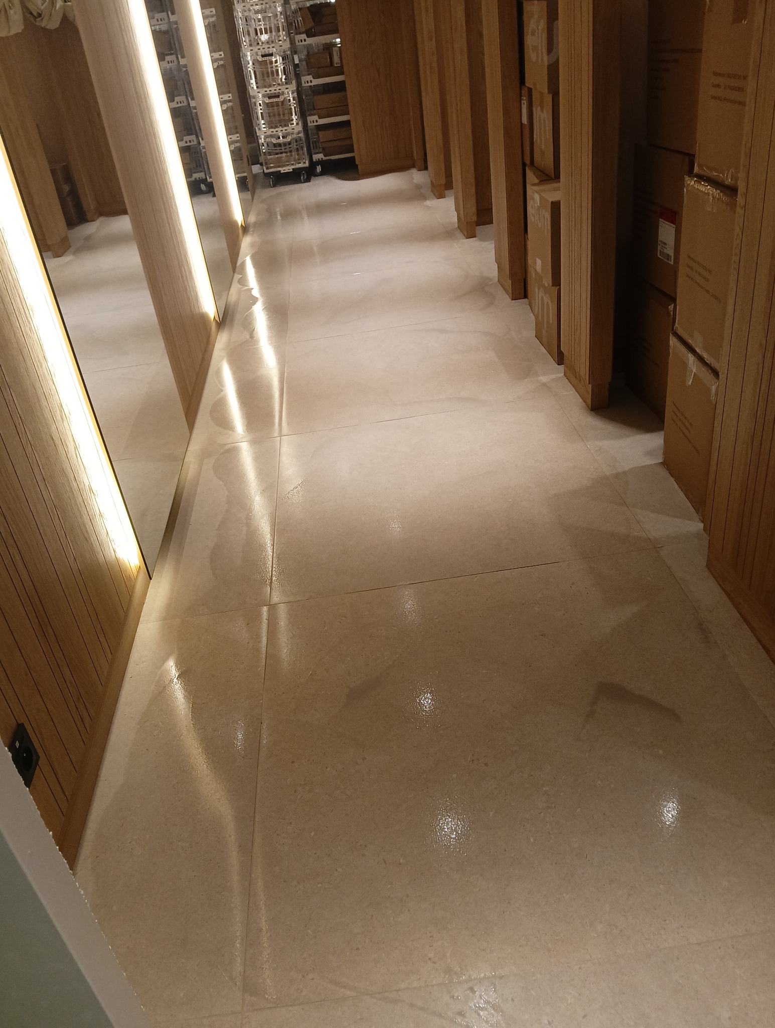 Błyszcząca, mokra podłoga z płytek w magazynie lub sklepie, oświetlona promieniami światła odbijającymi się od powierzchni, z widocznymi regałami i kartonami w tle.