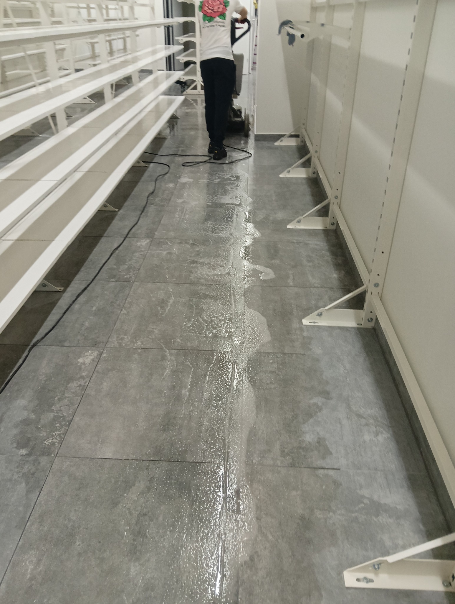 Czyszczenie posadzki w pomieszczeniu z regałami za pomocą szorowarki, widoczne mokre ślady po umytej powierzchni.