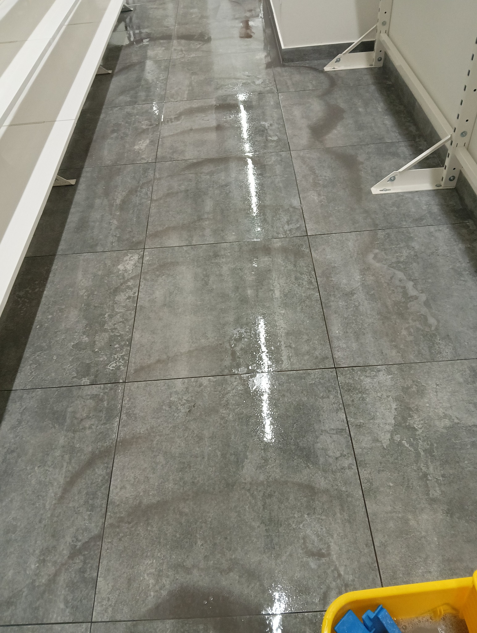 Świeżo umyta, mokra, szara podłoga z płytek w sklepie, z widocznymi białymi regałami po bokach i żółtym wiadrem z płynem do mycia w rogu.