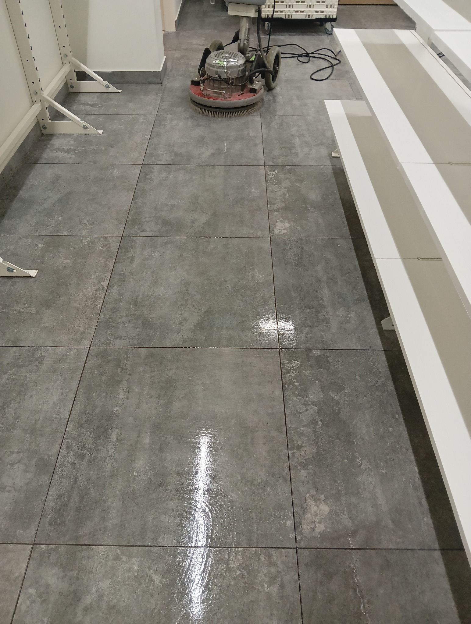 Automat do czyszczenia podłóg na szarych, kwadratowych płytkach, widoczny błyszczący ślad po czyszczeniu.