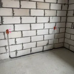 Instalacja elektryczna w surowym wnętrzu: pomarańczowe puszki podtynkowe z wystającymi przewodami na ścianie z białego bloczka, czarny peszel przy podłodze.