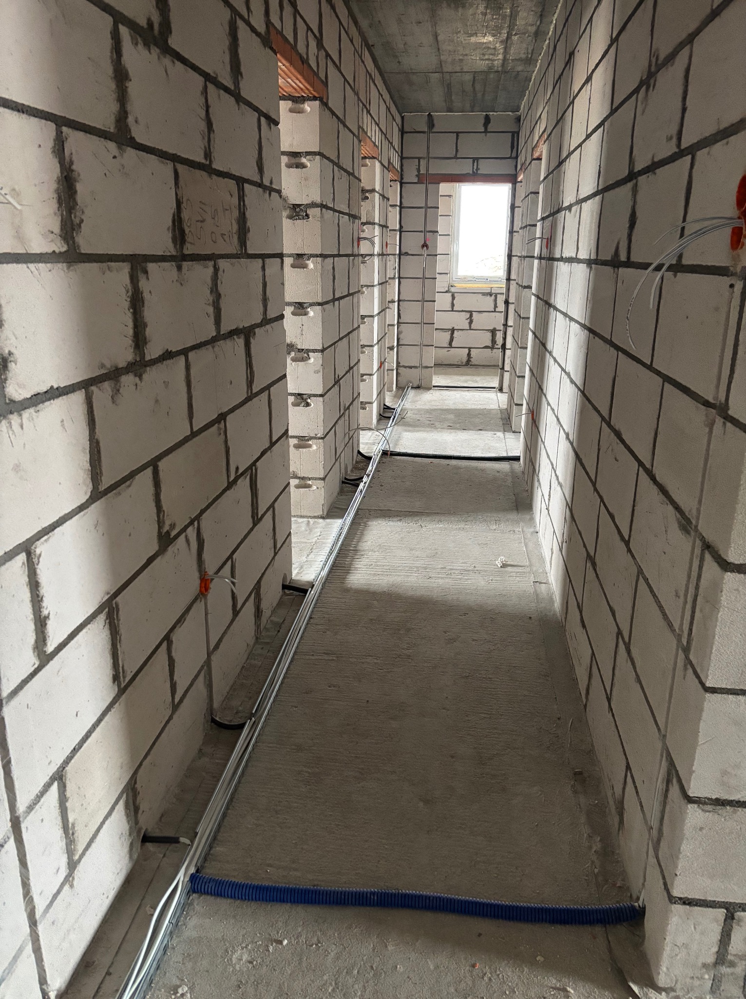 Korytarz w surowym stanie z widocznymi instalacjami elektrycznymi w metalowych korytkach i niebieskiej rurze karbowanej, ściany z białych bloczków, okno w oddali, widok w perspektywie.