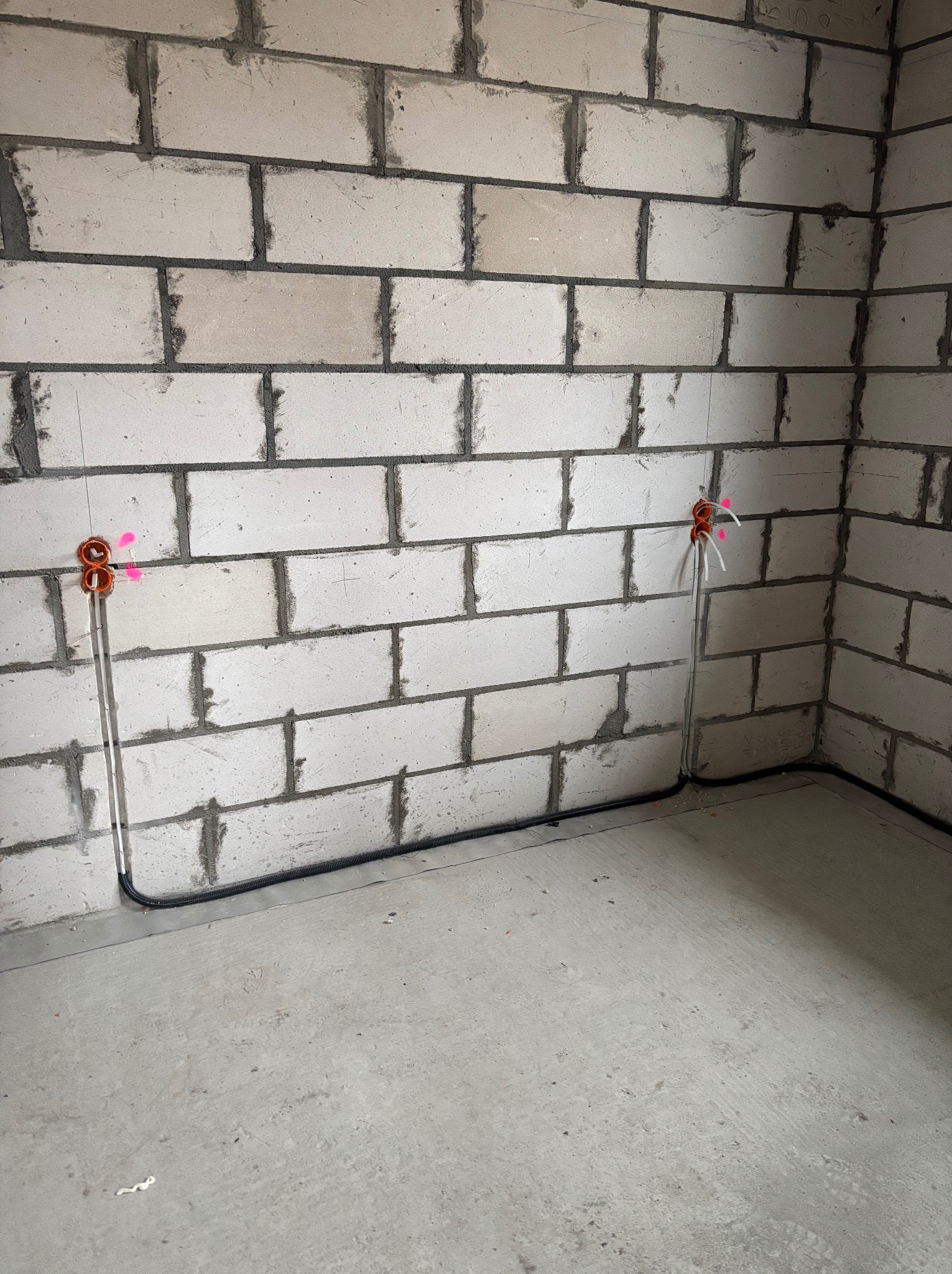 Instalacja elektryczna w surowym wnętrzu: pomarańczowe puszki podtynkowe z wystającymi przewodami na ścianie z białego bloczka, czarny peszel przy podłodze.