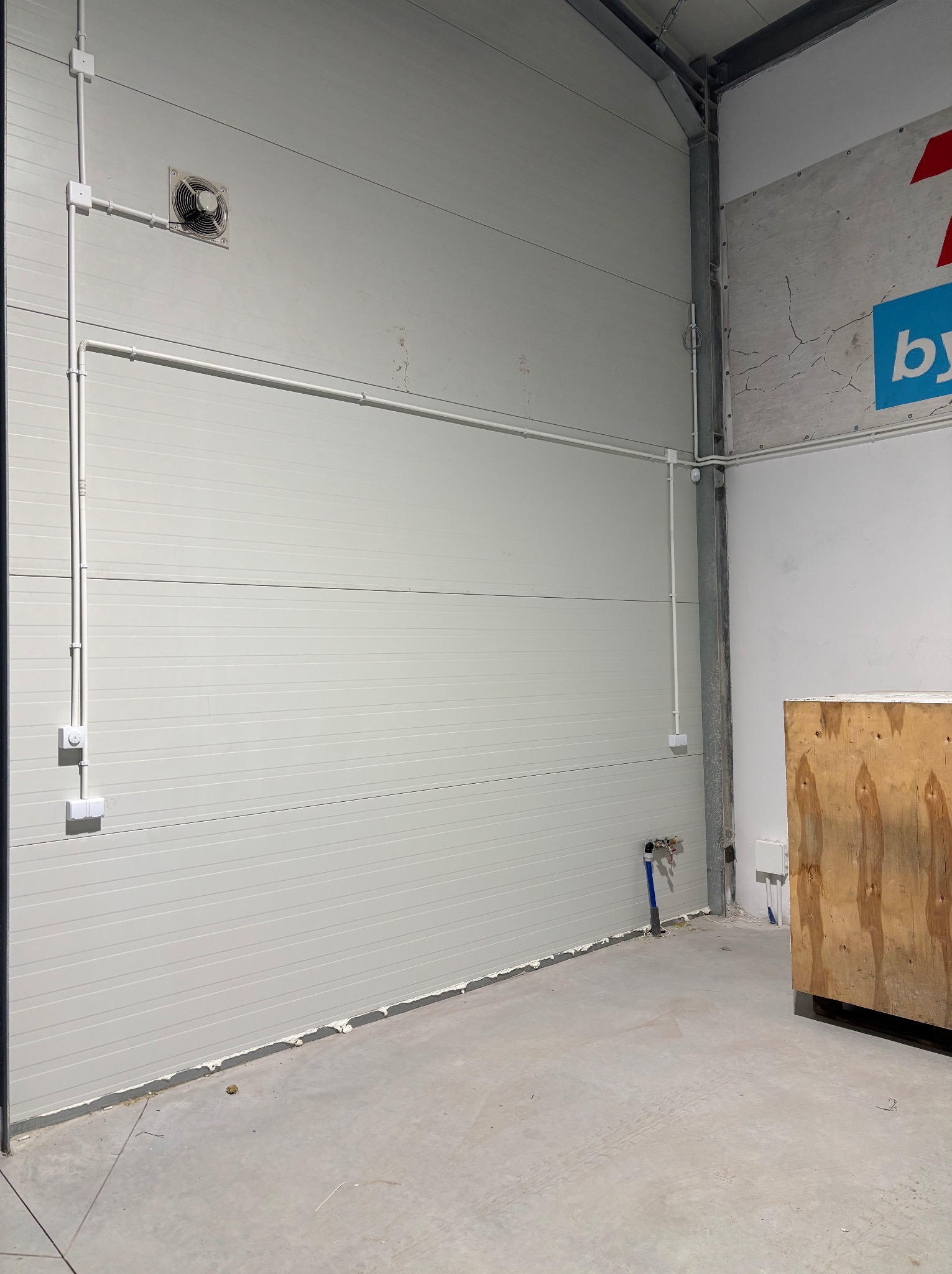 Wnętrze hali z szarą segmentową bramą, białe kable elektryczne w listwach, wentylator i fragment instalacji wodnej przy podłodze. Częściowo widoczny narożnik z betonową ścianą i fragmentem logo.