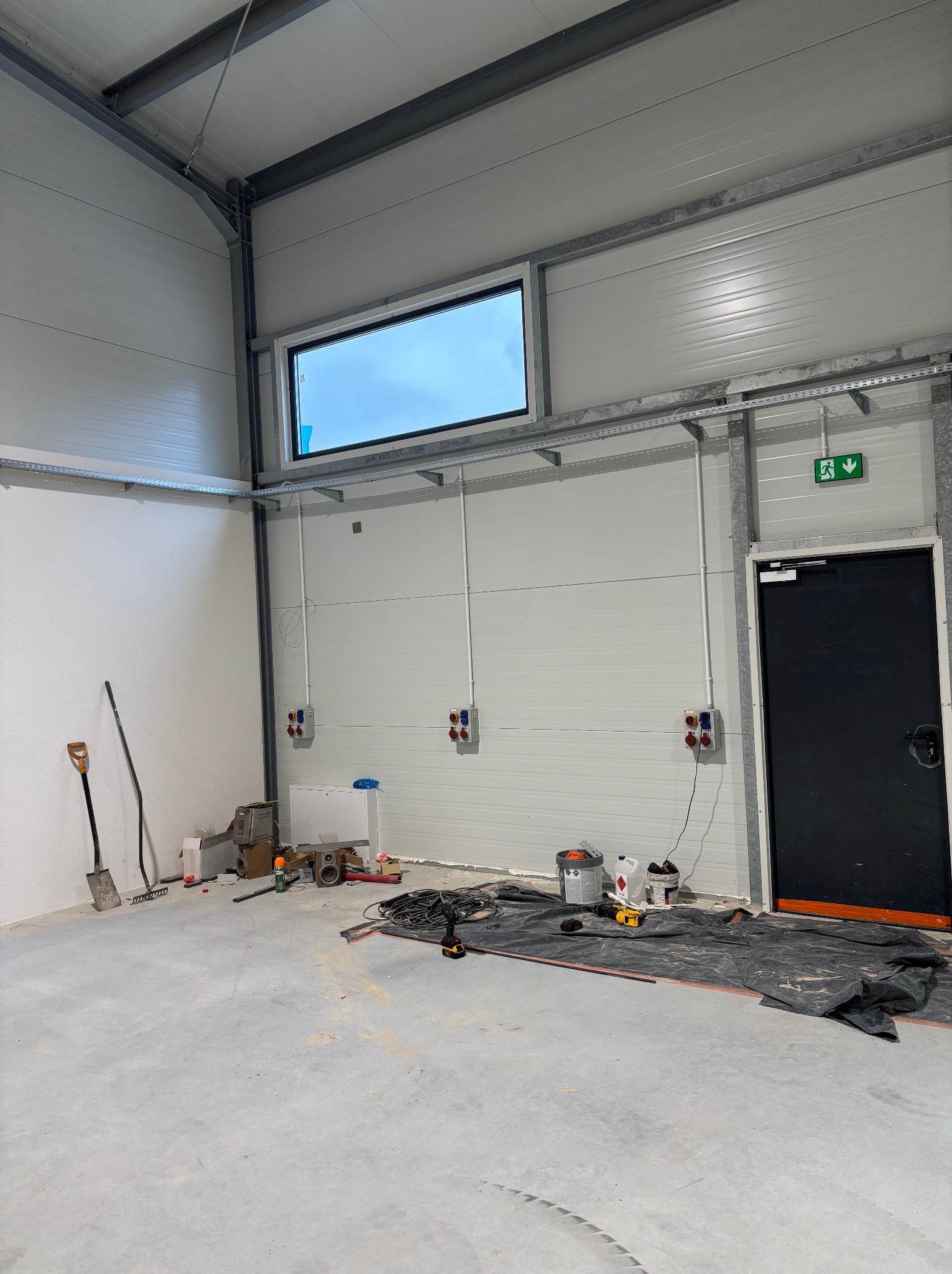 Wnętrze hali w trakcie prac: widoczne kable, puszki elektryczne na ścianie z panelami, narzędzia, szpachle, wiadro z farbą, czarna plandeka ochronna na podłodze, okno w górnej części ściany i znak...