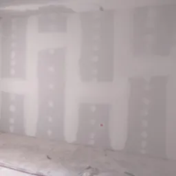 Ściana z płyt karton gipsowych po szpachlowaniu, widoczne łączenia i miejsca po wkrętach, szara podłoga w surowym stanie, metalowa konstrukcja przy ścianie.