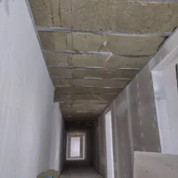 Korytarz w trakcie prac wykończeniowych: izolacja z wełny mineralnej zamontowana w metalowym stelażu pod sufitem, ściany przygotowane pod malowanie, widoczne przewody elektryczne i narzędzia.