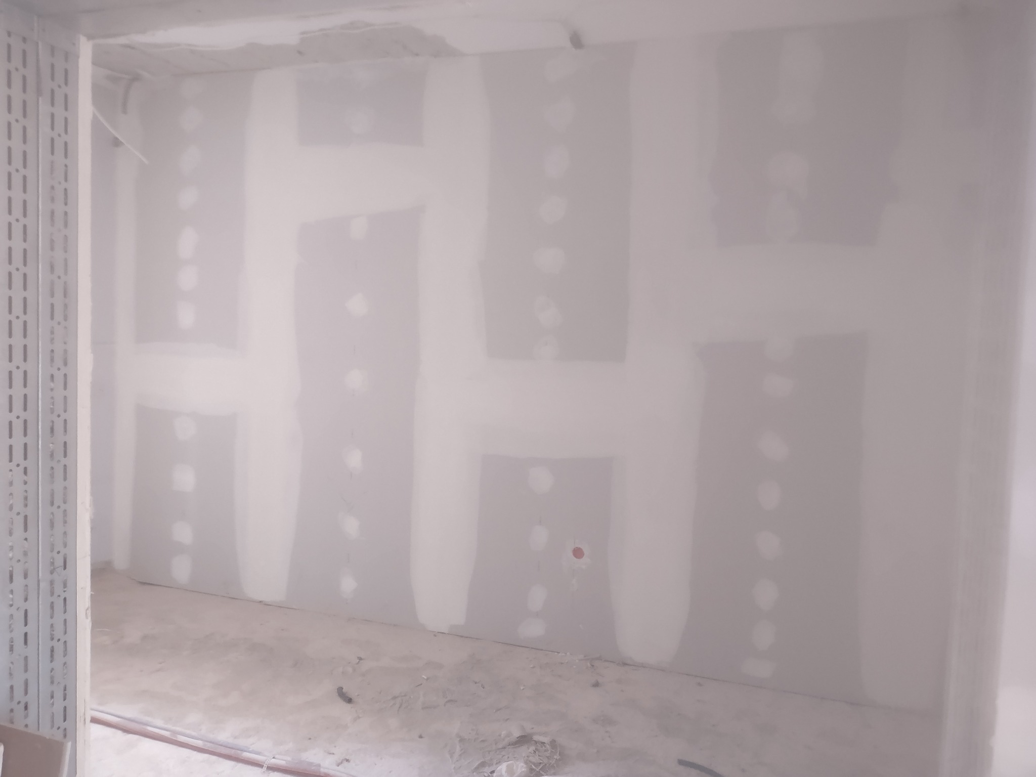 Ściana z płyt karton gipsowych po szpachlowaniu, widoczne łączenia i miejsca po wkrętach, szara podłoga w surowym stanie, metalowa konstrukcja przy ścianie.