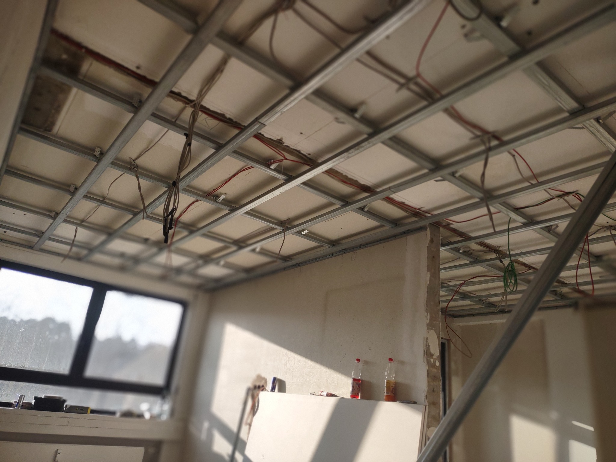 Stelaż sufitowy z metalowych profili w trakcie montażu, widoczne przewody elektryczne, ściany w stanie surowym, butelki z napojami na parapecie.