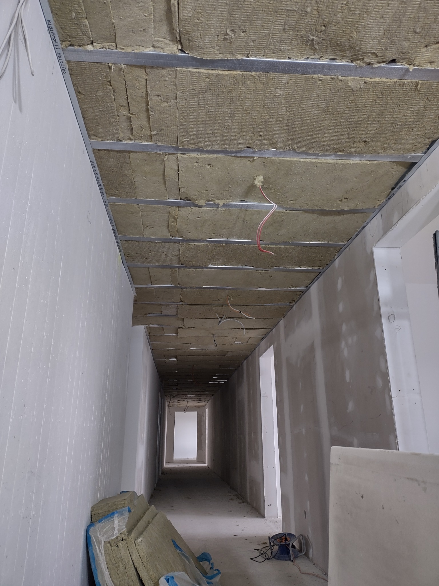 Korytarz w trakcie prac wykończeniowych: izolacja z wełny mineralnej zamontowana w metalowym stelażu pod sufitem, ściany przygotowane pod malowanie, widoczne przewody elektryczne i narzędzia.