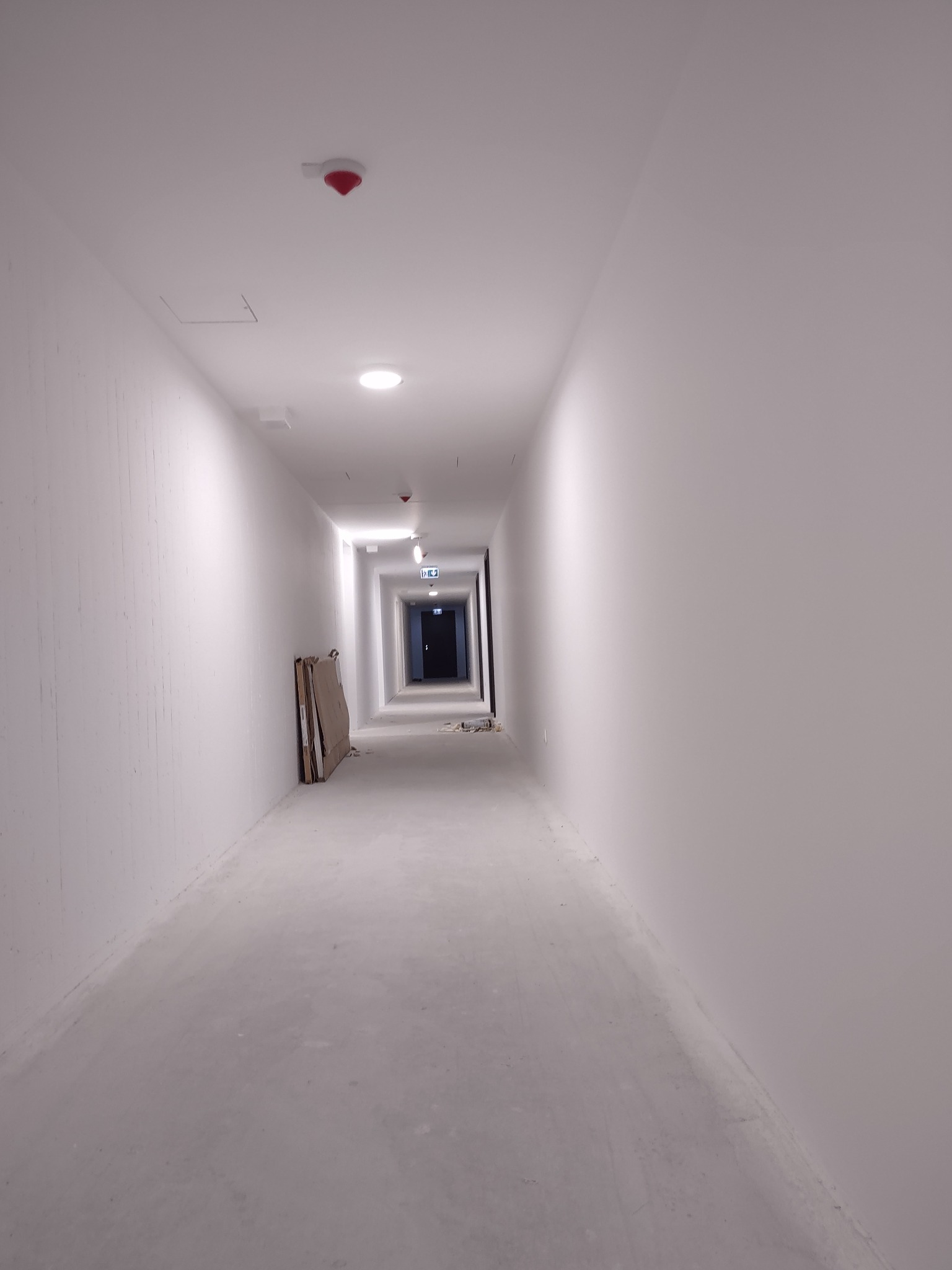 Długi korytarz w stanie surowym z widocznymi śladami po pracach wykończeniowych, lekko szare ściany i podłoga, na ścianie widoczne płyty kartonowo-gipsowe przed obróbką.