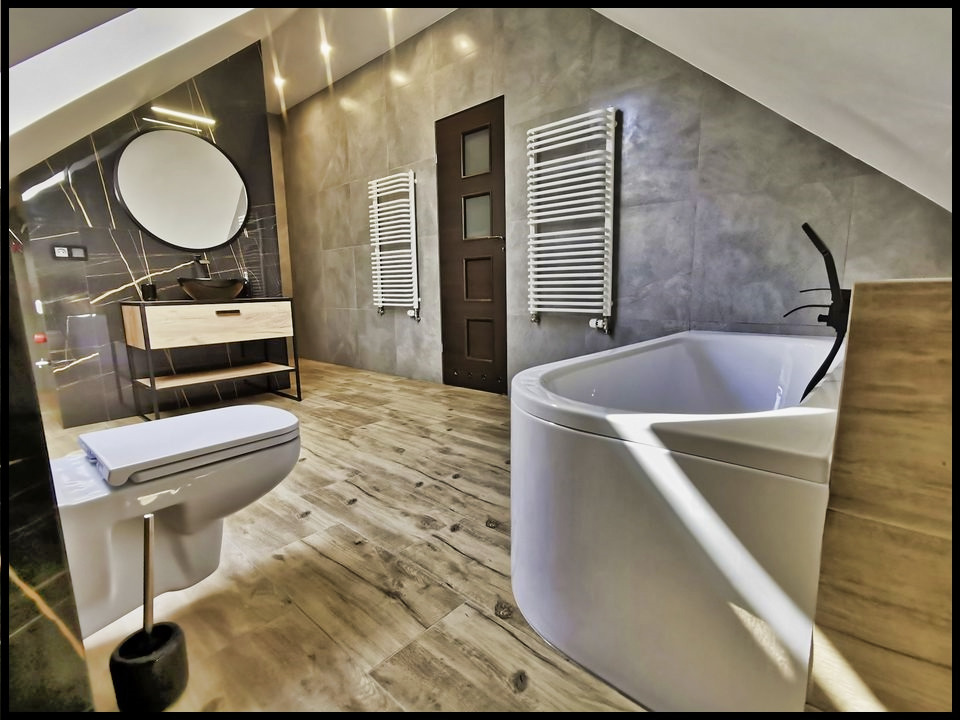 Nowoczesna łazienka na poddaszu z drewnopodobną podłogą, owalnym lustrem z podświetleniem LED, umywalką nablatową, grzejnikami drabinkowymi i białą wanną narożną.