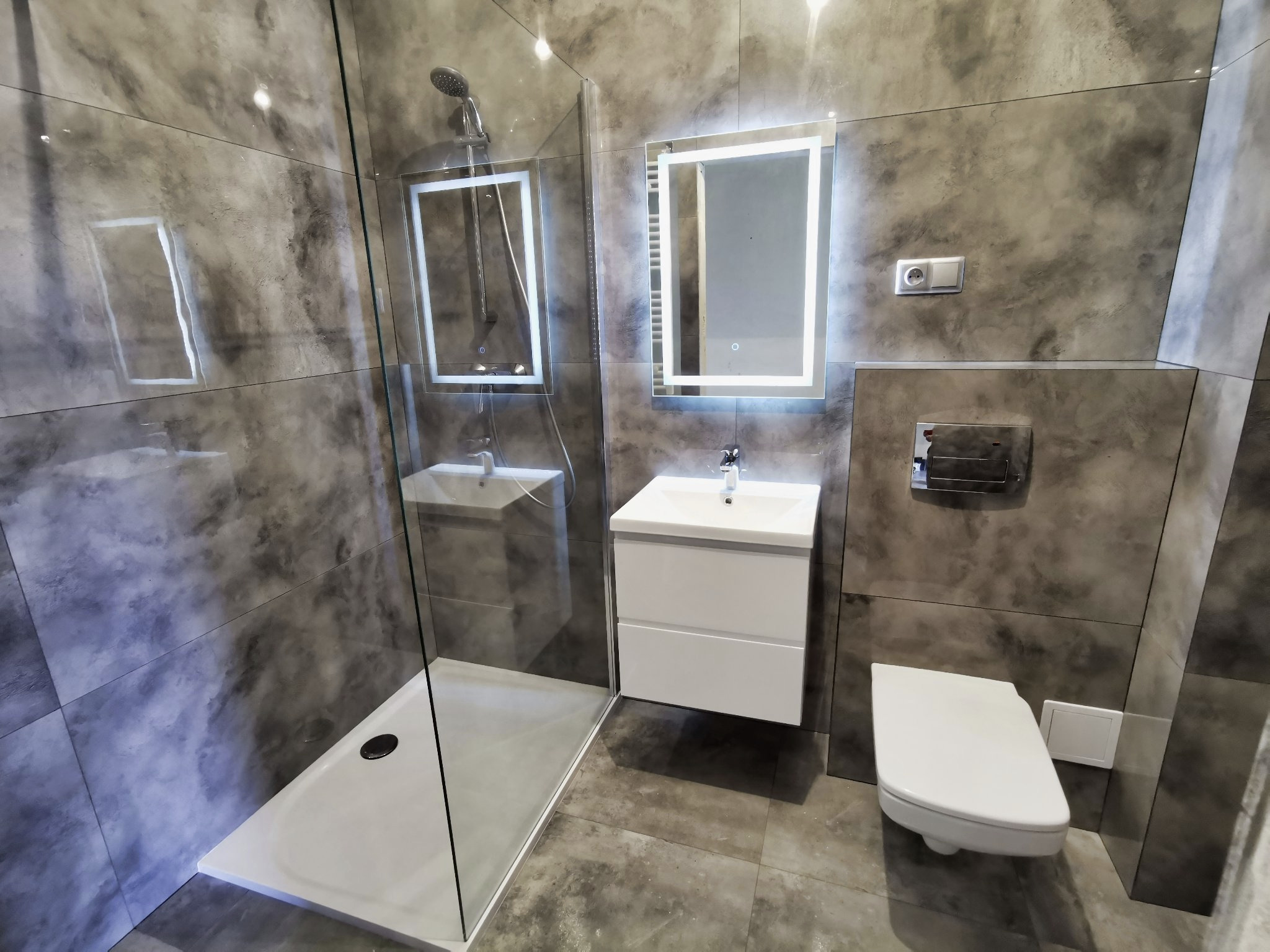 Nowoczesna łazienka z kabiną prysznicową typu walk-in, podświetlanym lustrem i wiszącą toaletą, wykończona płytkami imitującymi beton.