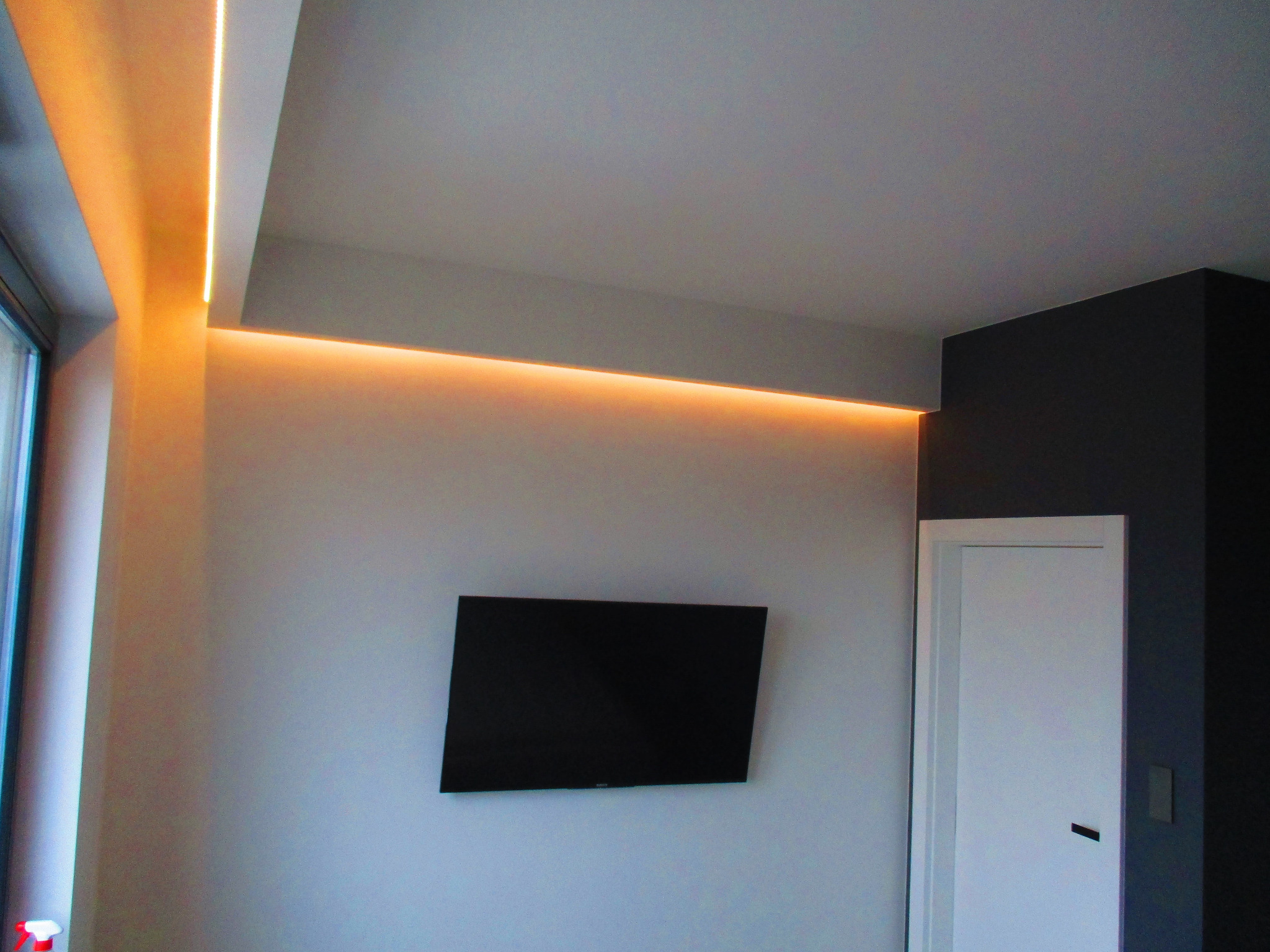 Nowoczesne oświetlenie LED w suficie podwieszanym, ciepła barwa światła, szara ściana z telewizorem i białe drzwi w minimalistycznym wnętrzu.