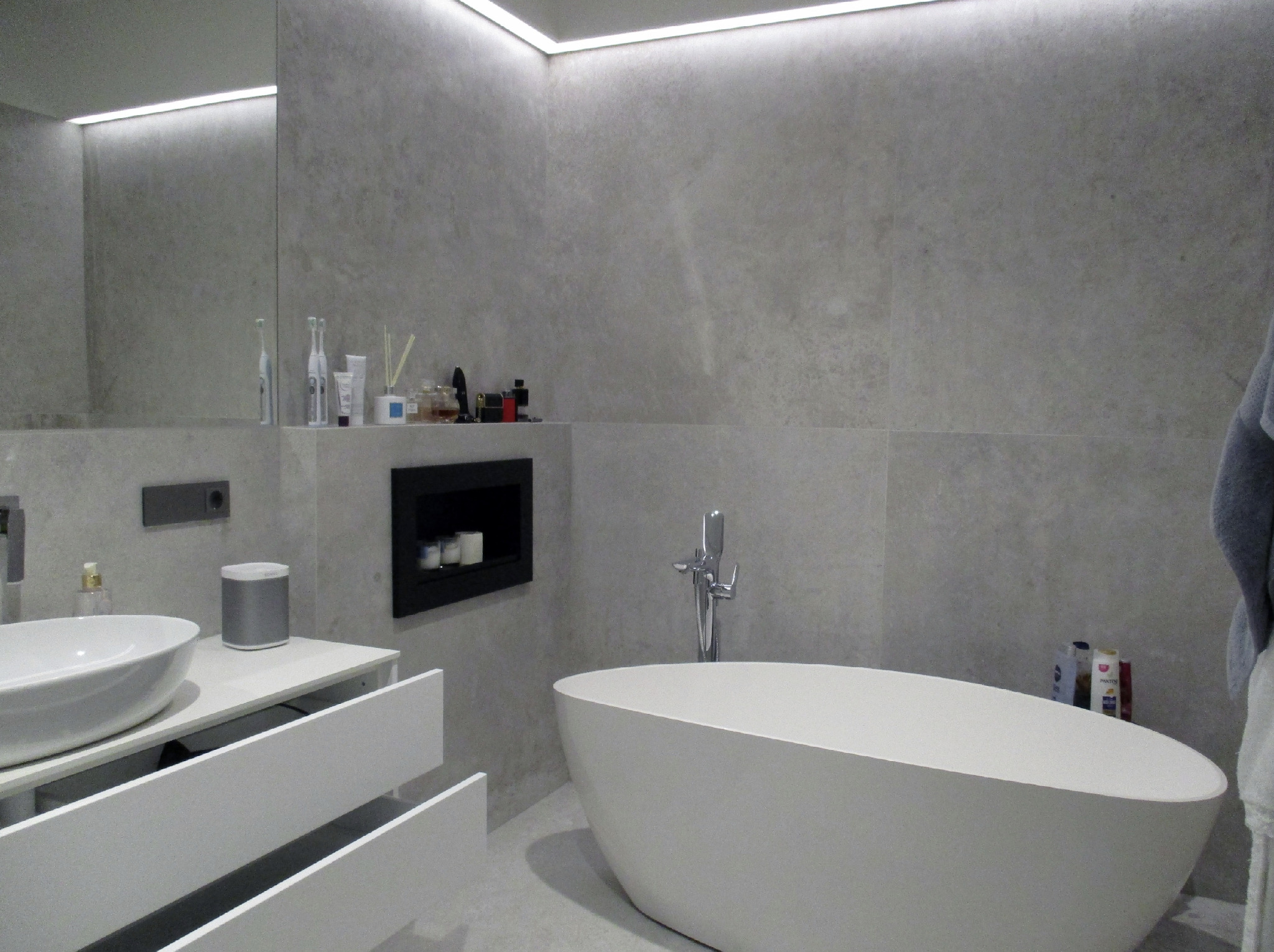 Nowoczesna łazienka w odcieniach szarości z wolnostojącą białą wanną, umywalką nablatową na szafce z szufladami i wnęką z dekoracyjnymi świecami.